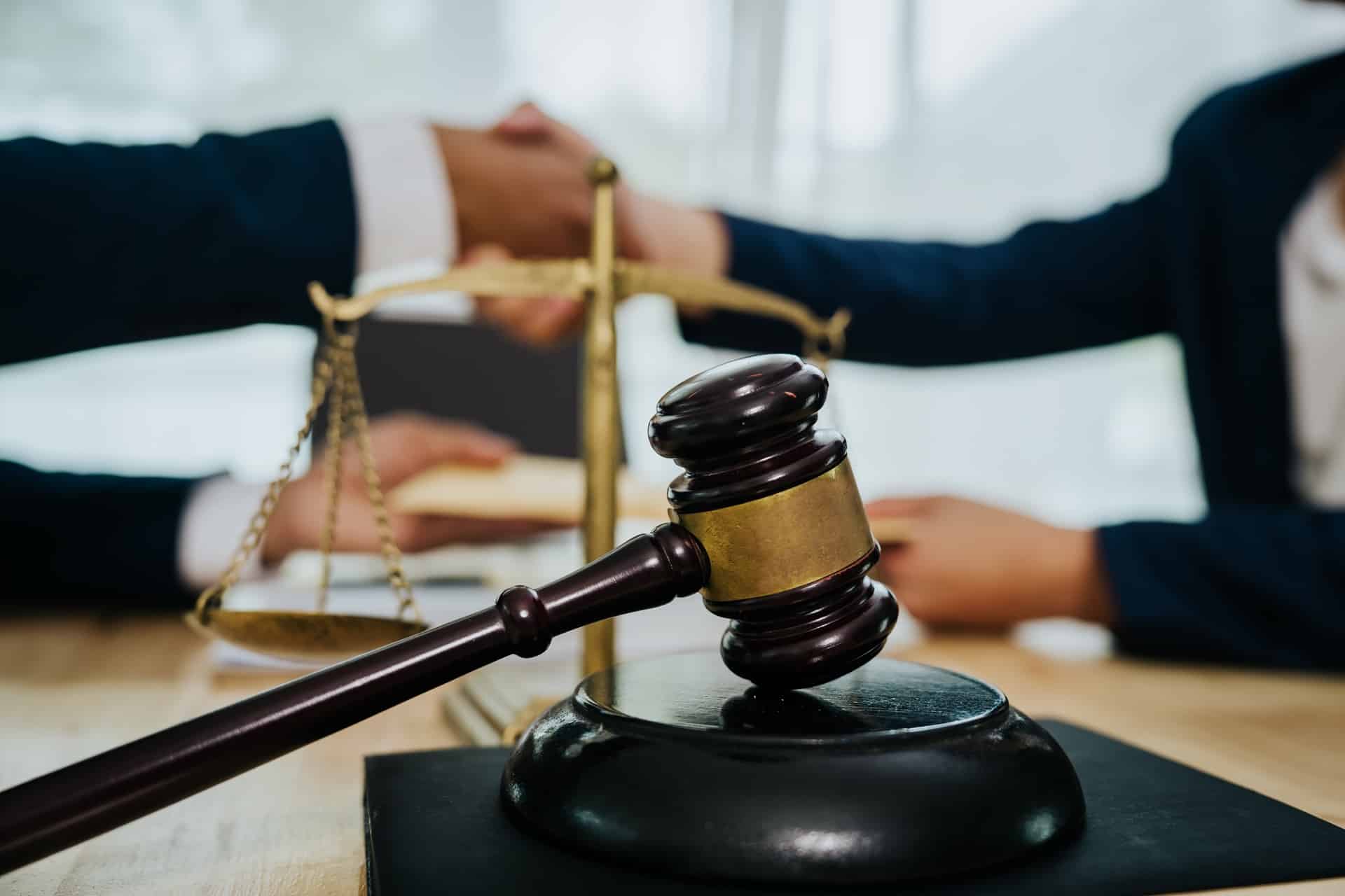 A lawyer shakes hands with a client at a desk in front of scales of justice and a gavel resting on its stand.