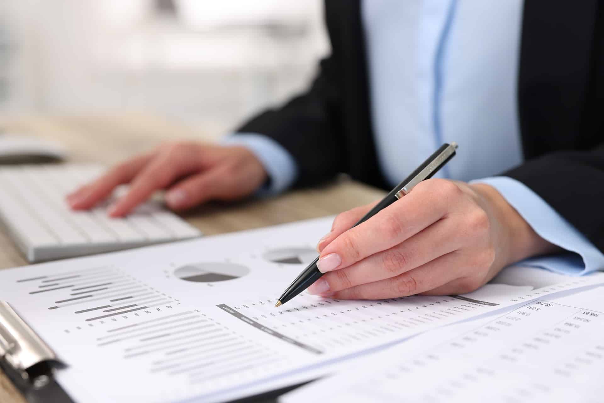 A person sitting at a desk with one hand holding a pen on some paperwork and the other resting on a white keyboard.