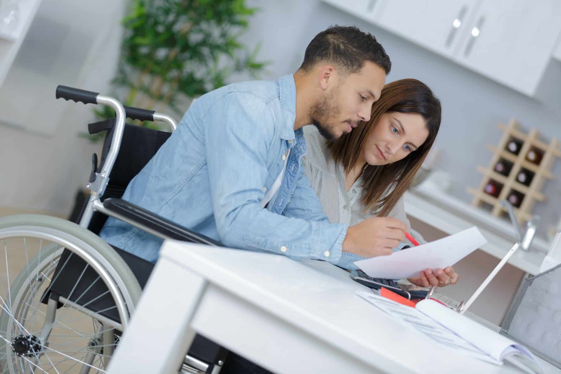One person in a wheelchair sitting next to a woman sitting at a desk filling out paperwork